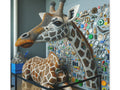 A canvas art piece depicting a three-dimensional giraffe protruding from the canvas, embellished with an intricate mosaic of various buttons and beads against a background of neatly organized craft materials.