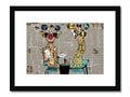 Two giraffe standing on the side of a field together behind an art print of their